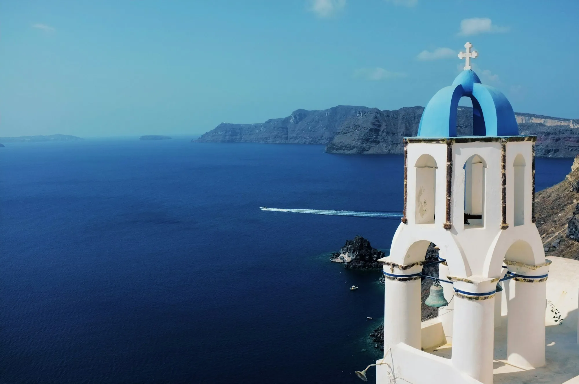 Campanário de cúpula azul em Santorini sobre a caldeira, com barco cruzando o mar Egeu